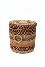 Panier artisanal africain tressé vu de profil, couvercle légèrement soulevé — bandes horizontales multicolores en raphia naturel, orange et bordeaux, anse latérale visible, fond blanc