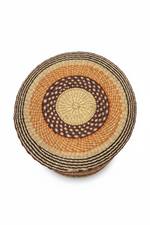 Panier artisanal africain tressé vu de dessus — couvercle rond avec motif spirale concentrique en raphia naturel, orange et brun foncé, fond blanc

