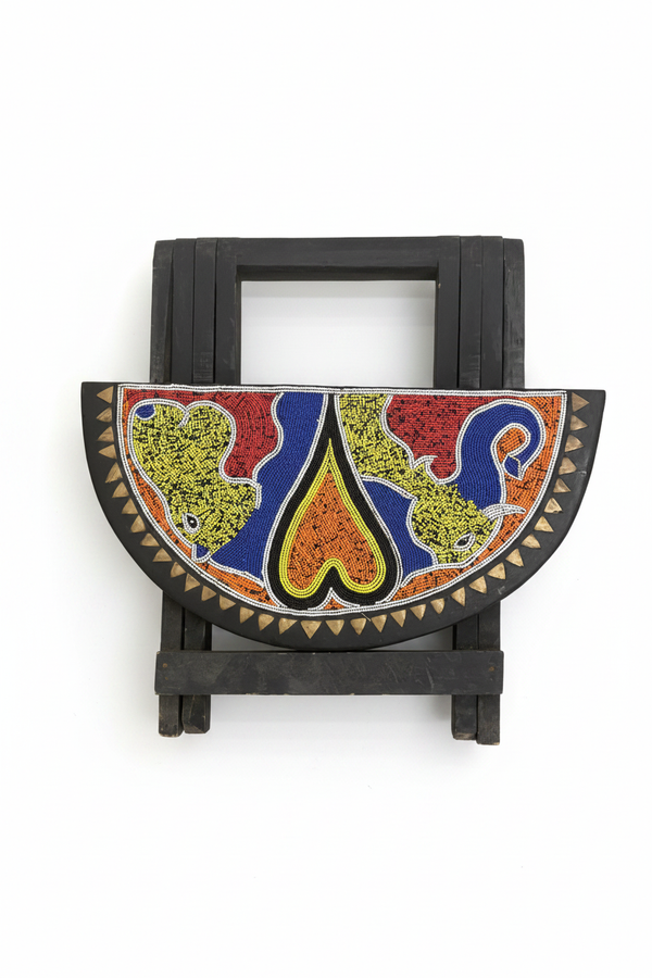 Tabouret pliant africain artisanal en bois noir avec demi-cercle perlé à motifs d'animaux en vert olive, bleu cobalt et orange  forme centrale en goutte jaune-orangée, bordure de triangles en métal doré. Fond blanc studio.
